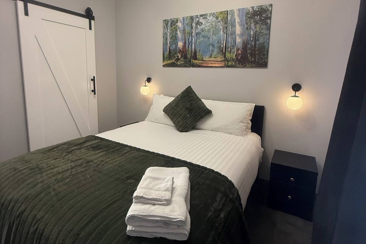 Ein Schlafzimmer mit einem Bett, einem Bild, zwei Nachttischlampen, einer Tür und einem Nachttisch.