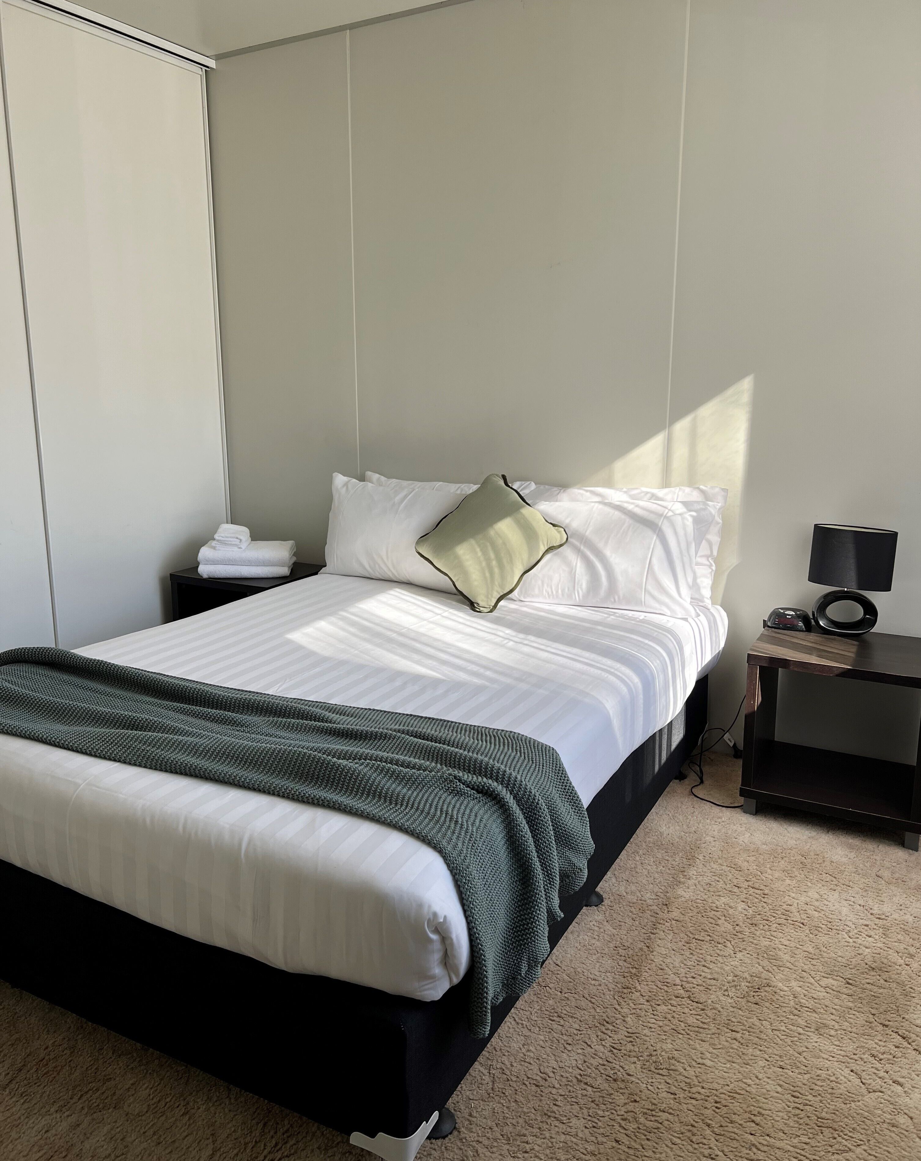 A neatly made bed with white linens and a green throw blanket, a bedside table with a lamp, and a wall-mounted phone.