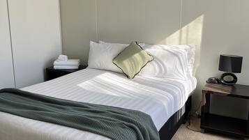 A neatly made bed with white linens and a green throw blanket, a bedside table with a lamp, and a wall-mounted phone.