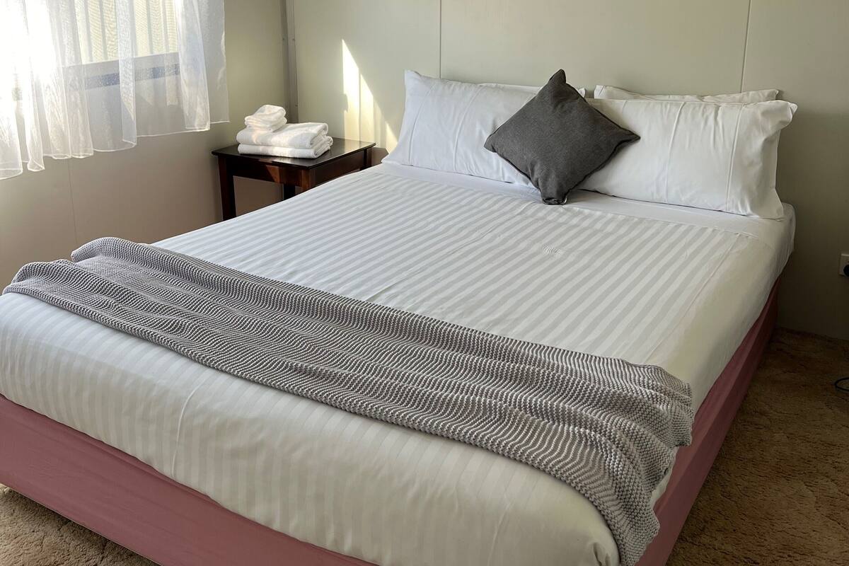 Ein ordentlich gemachtes Bett mit rosa Basis, weißen Leinen und einer gestreiften Decke. Ein Nachttisch mit einer weißen Lampe und einem gefalteten Handtuch steht daneben. Ein durchsichtiger weißer Vorhang bedeckt teilweise ein Fenster.