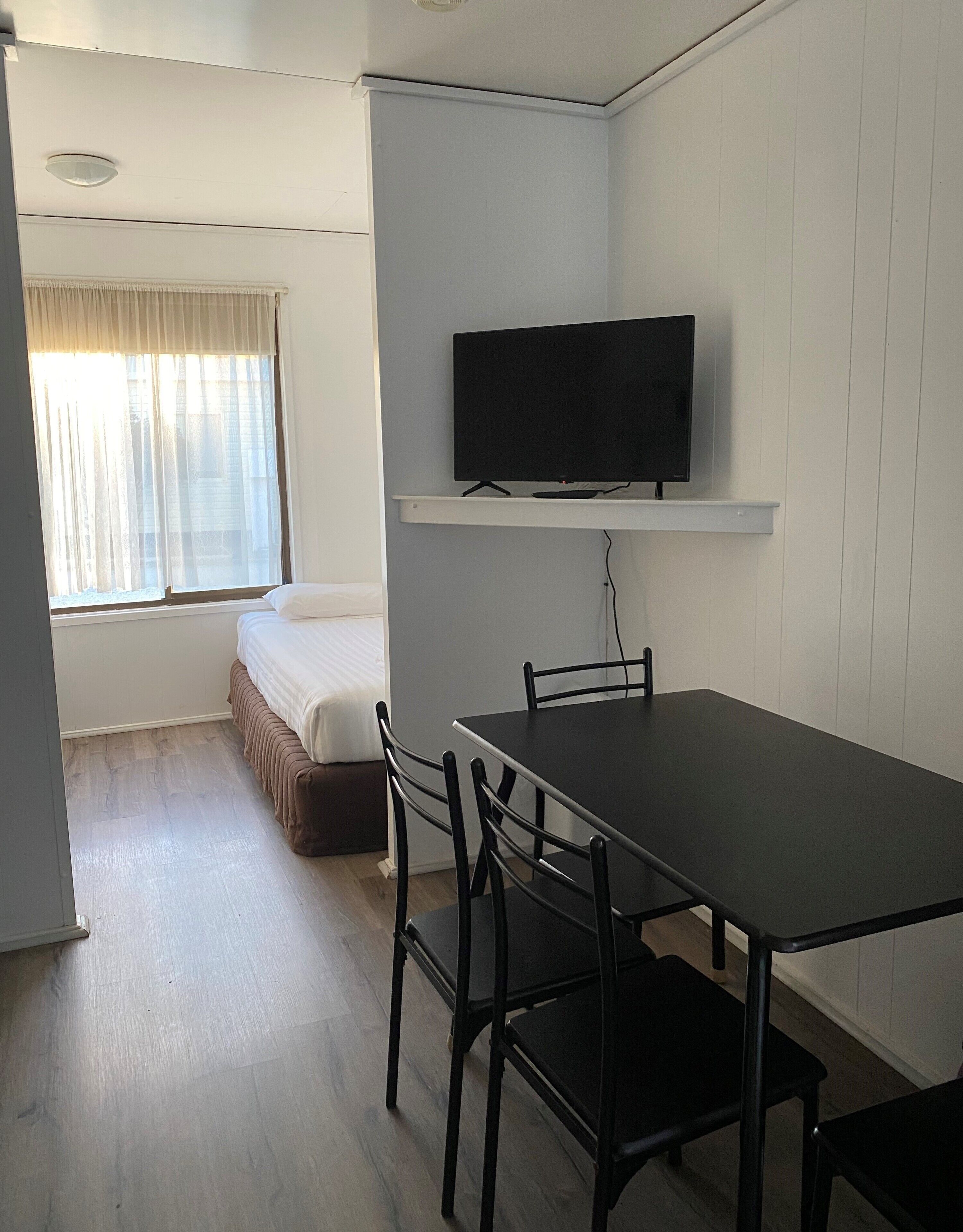 A small, clean room with a bed, a TV mounted on a wall, a table with chairs, and a window with curtains.
