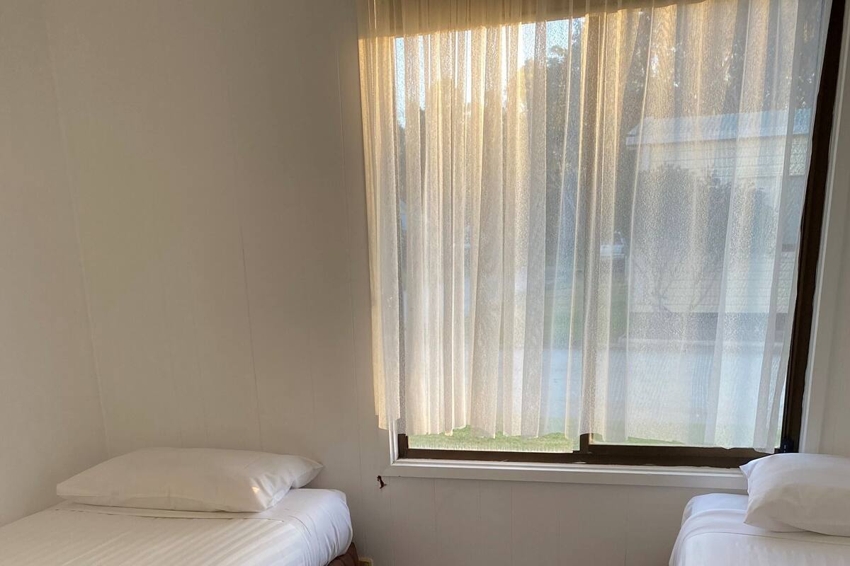 Ein Zimmer mit zwei Betten, Holzboden, einem Fenster mit durchsichtigen Vorhängen und Blick auf ein Gebäude.