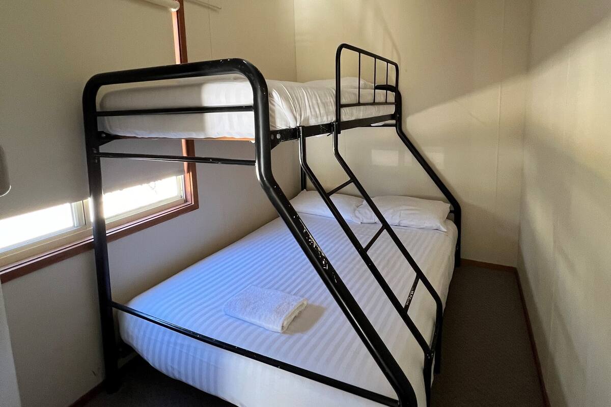 Ein Etagenbett in einem kleinen Zimmer mit Fenster, Teppichboden und einer sichtbaren Tür.