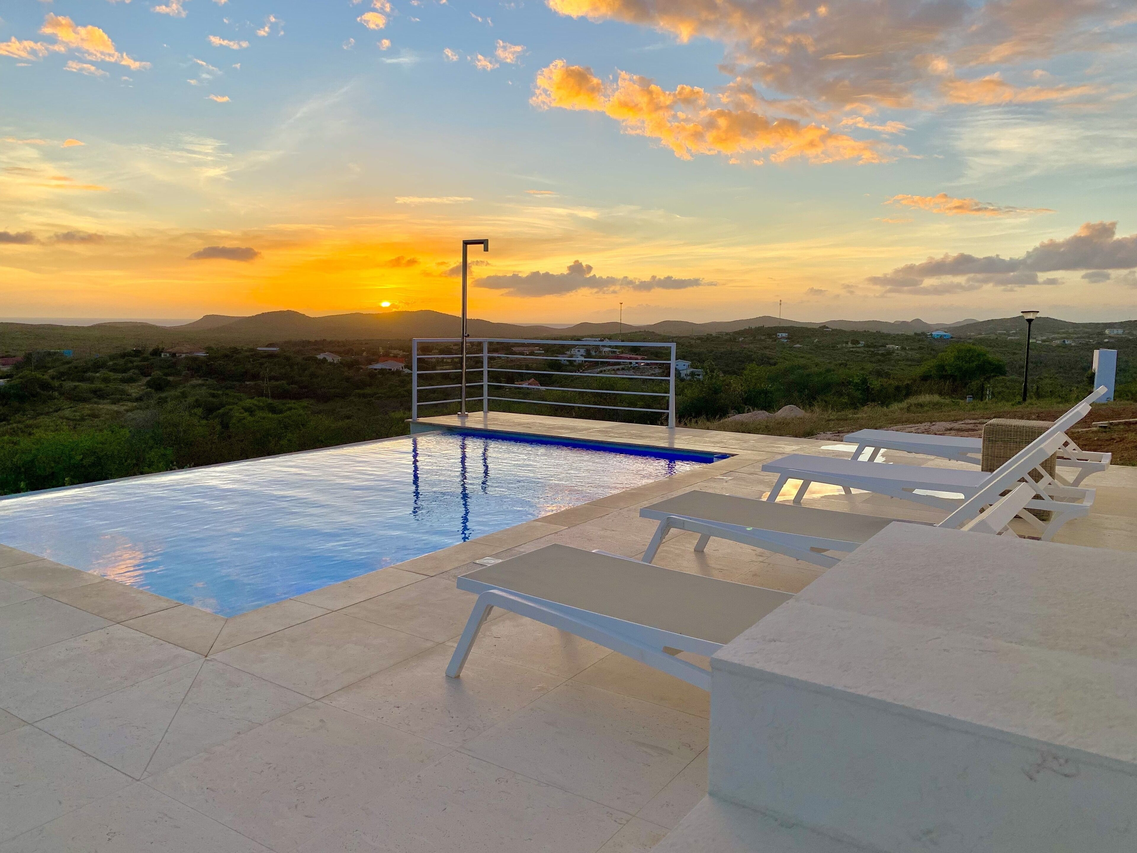 Una piscina con barandilla metálica, rodeada de sillas de descanso y vista al atardecer sobre un paisaje montañoso.