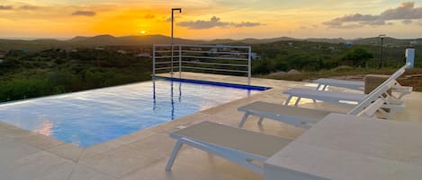 Una piscina con barandilla metálica, rodeada de sillas de descanso y vista al atardecer sobre un paisaje montañoso.