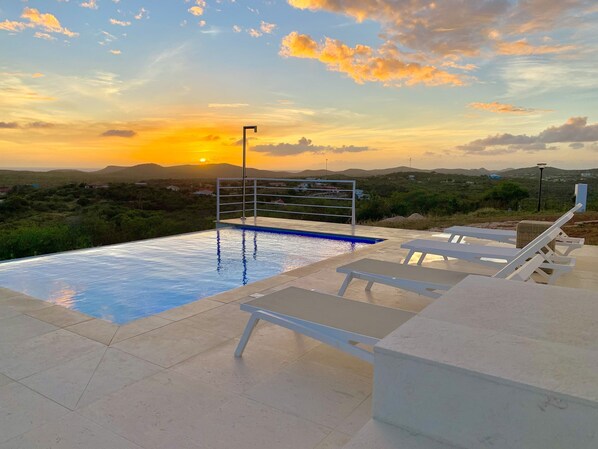 A swimming pool with a metal railing, surrounded by lounge chairs and a view of the sunset over a hilly landscape.