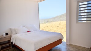 A bedroom with a bed, a nightstand, a window with blinds, and a view of a desert landscape. - FonteinTop - 360° Panoramic View (Willemstad)