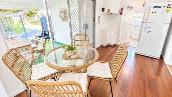 Uma área de refeições com cadeiras de vime e mesa de vidro, uma cozinha com armários brancos e vista para o exterior.