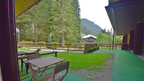 Una terrazza esterna con tavolo e sedie in legno, prato sintetico verde e vista su una montagna e una foresta sullo sfondo.