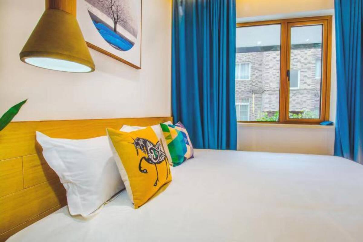 Een hotelkamer met een bed, een raam met blauwe gordijnen, een schilderij aan de muur en een lamp.