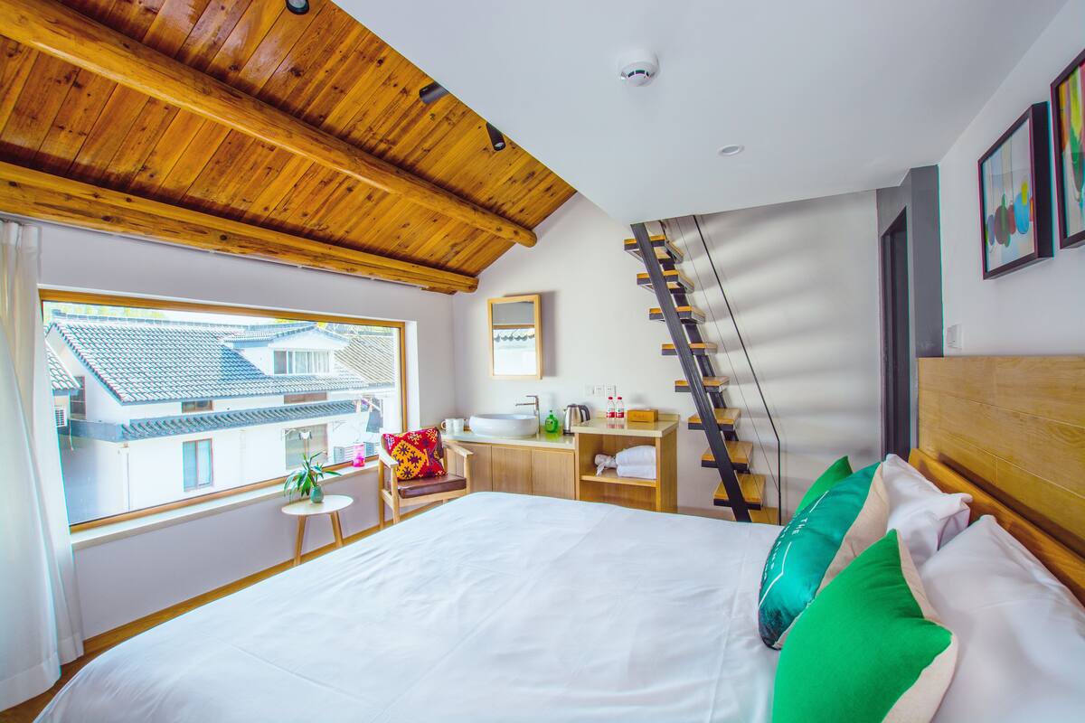 Een slaapkamer met een groot bed, houten plafond, raam met uitzicht op een traditioneel gebouw, een klein tafeltje met een plant, en een badkamer met wastafel en spiegel.
