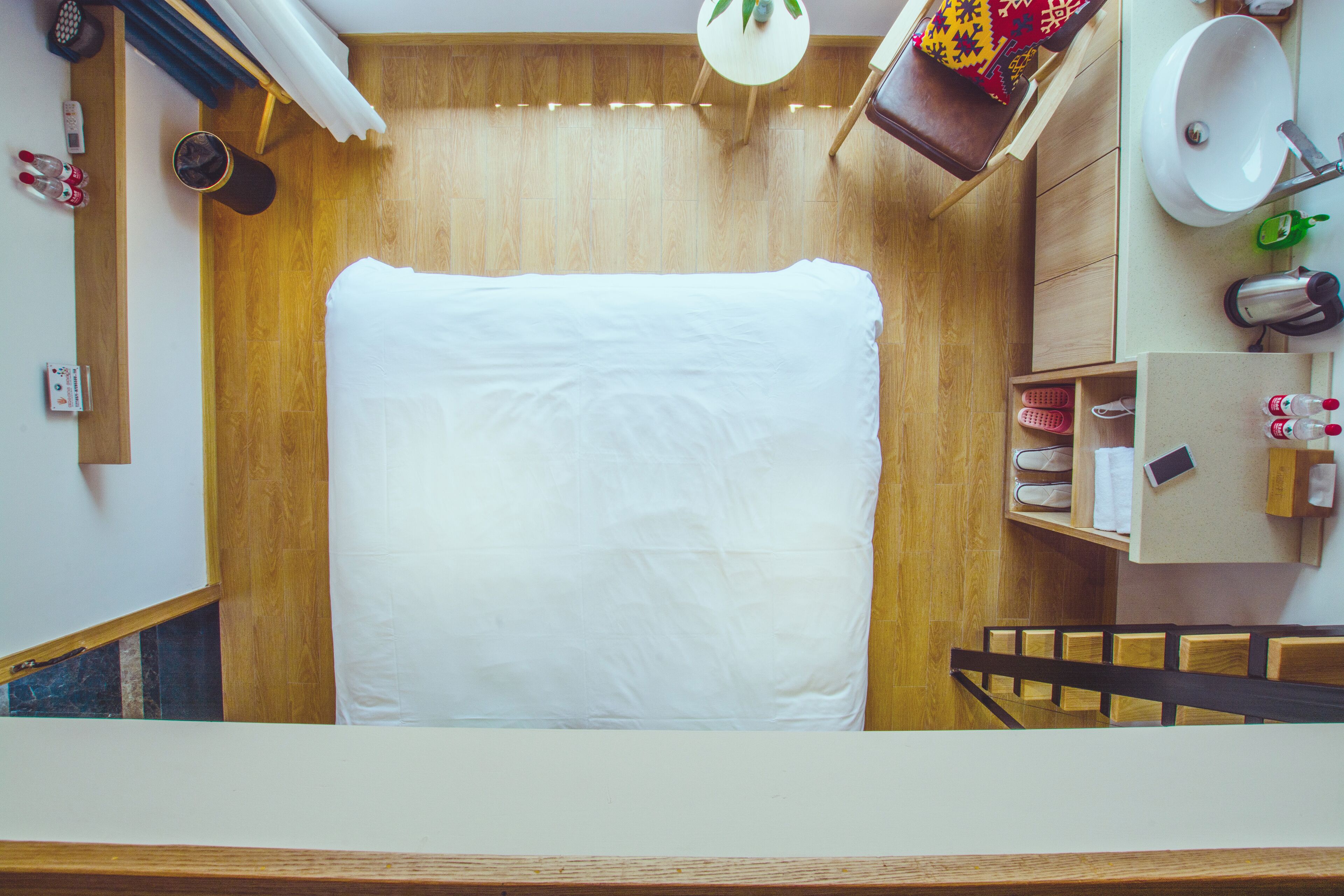 Un dormitorio pequeño con cama, suelo de madera, un baño compacto con lavamanos y espejo, y una escalera que sube a otro nivel.