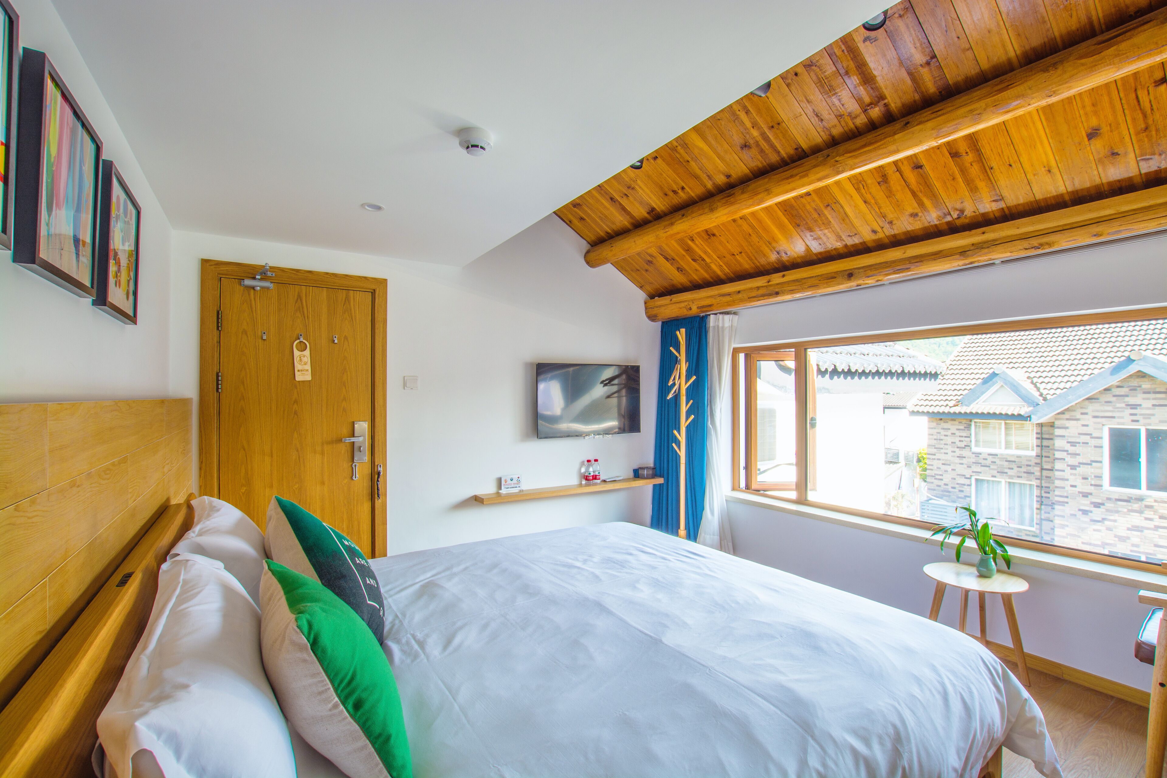 A bedroom with a wooden ceiling, a bed with white and green pillows, a TV on the wall, and a window with a view of buildings.