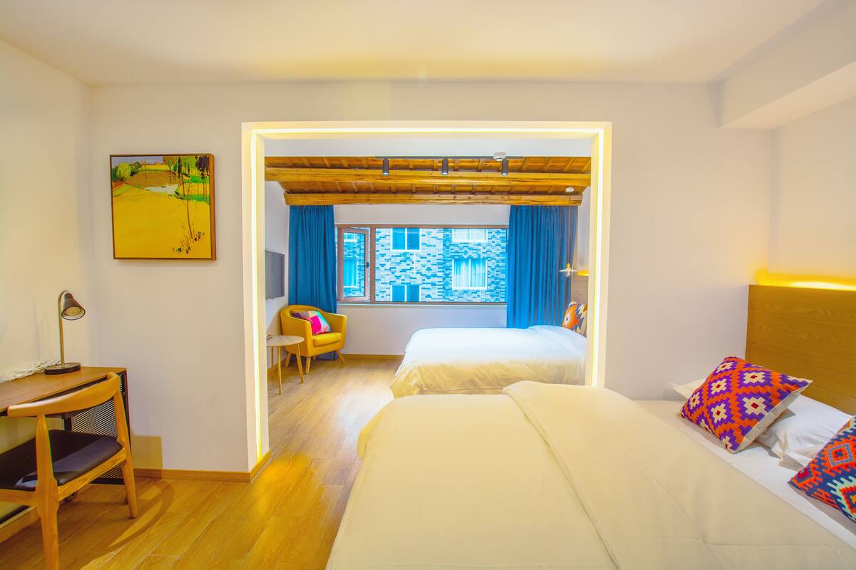 Hotelkamer met een houten bureau, een stoel, twee bedden, een kleurrijk schilderij en uitzicht op een gebouw door een raam met blauwe gordijnen.