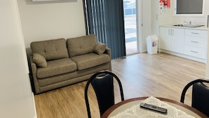 Una sala de estar con un sofá, una mesa de comedor con sillas, una zona de cocina con lavaplatos y armarios, y un ventanal con cortinas.