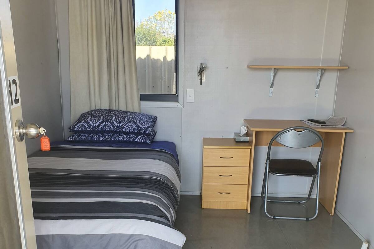 A compact hotel room with a bed, a desk, a chair, a small cabinet, and a window with a view of greenery.