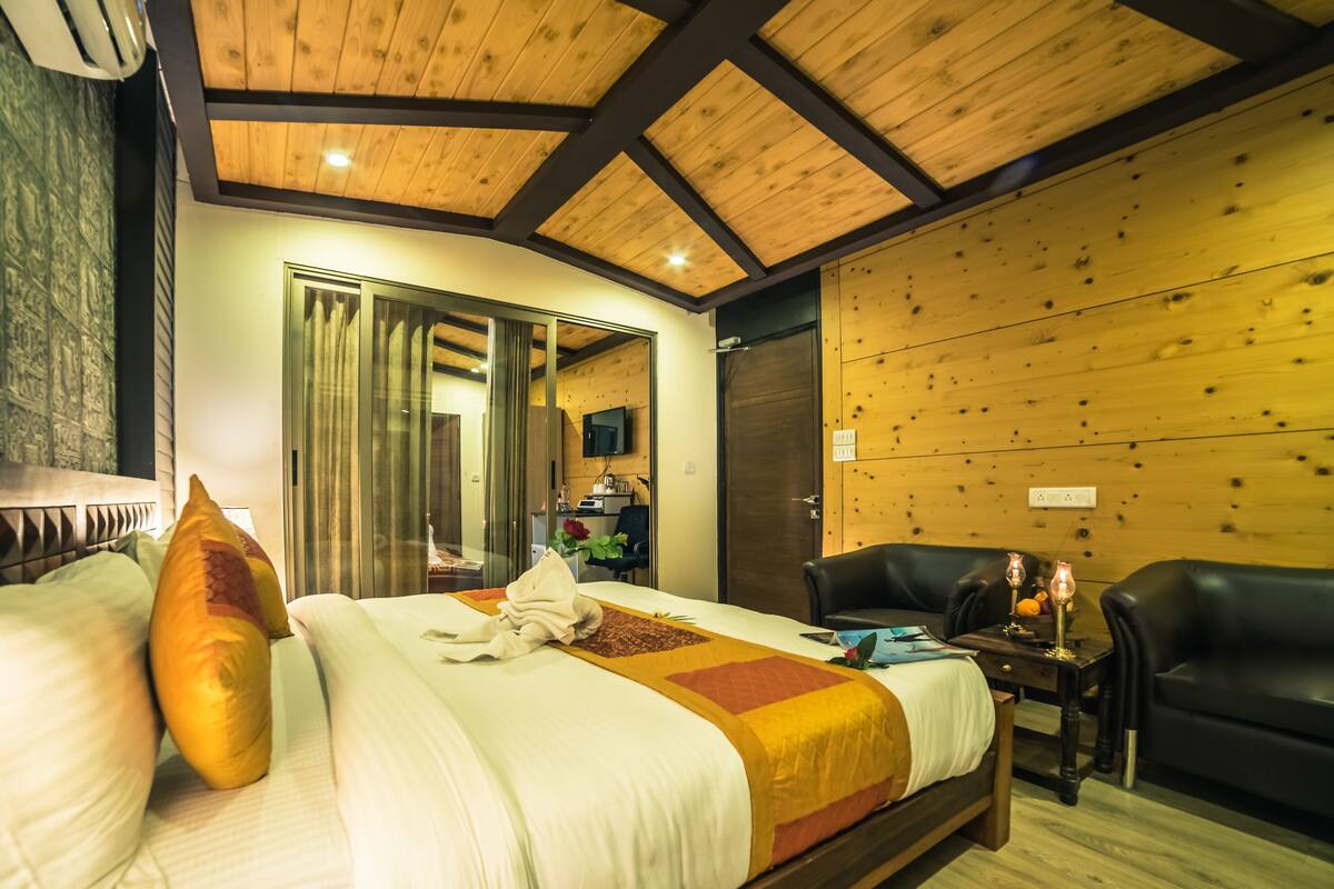 Een hotelkamer met een houten plafond, een groot bed met een wit-oranje sprei, een zwarte leren fauteuil en een kleine tafel met een vaas en kaarsen.