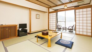 Una habitación japonesa tradicional con suelo de tatami, una mesa de madera baja, un televisor y una puerta corredera que da a un balcón.
