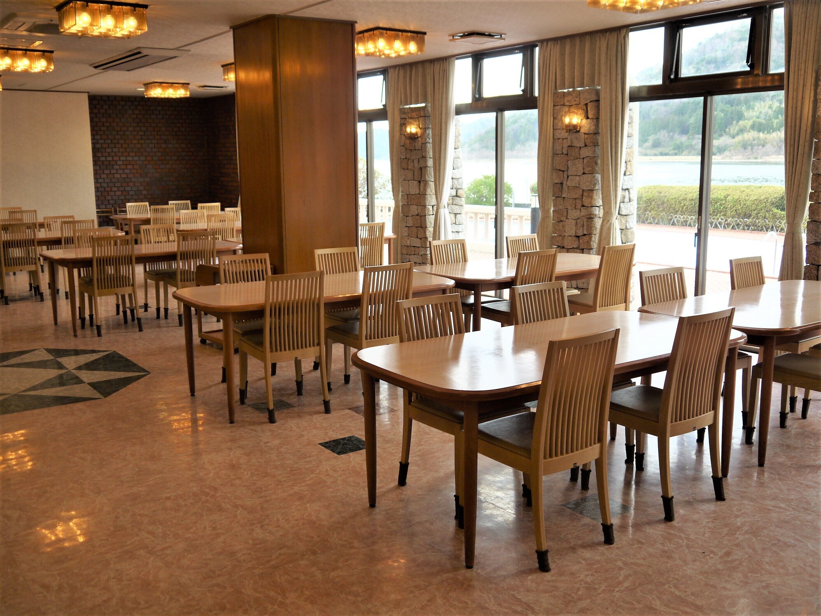 A dining area with multiple tables and chairs, large windows with curtains, and a stone wall.