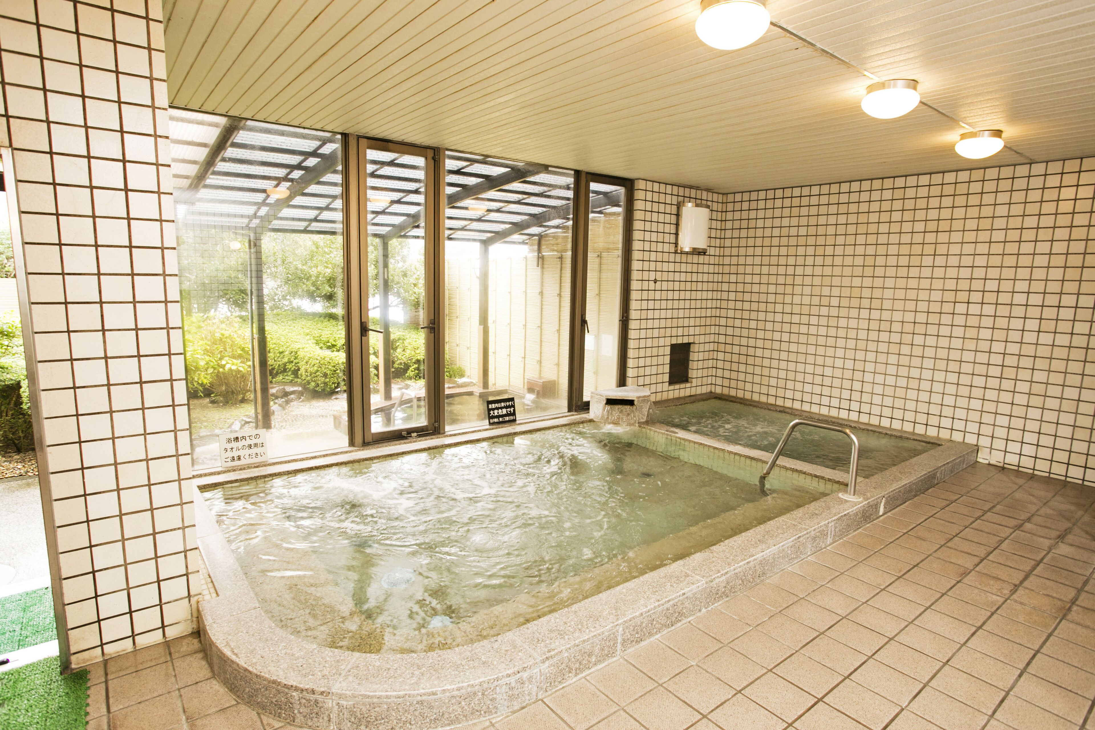 A hot tub in a room with tiled walls and a glass door leading to a garden.