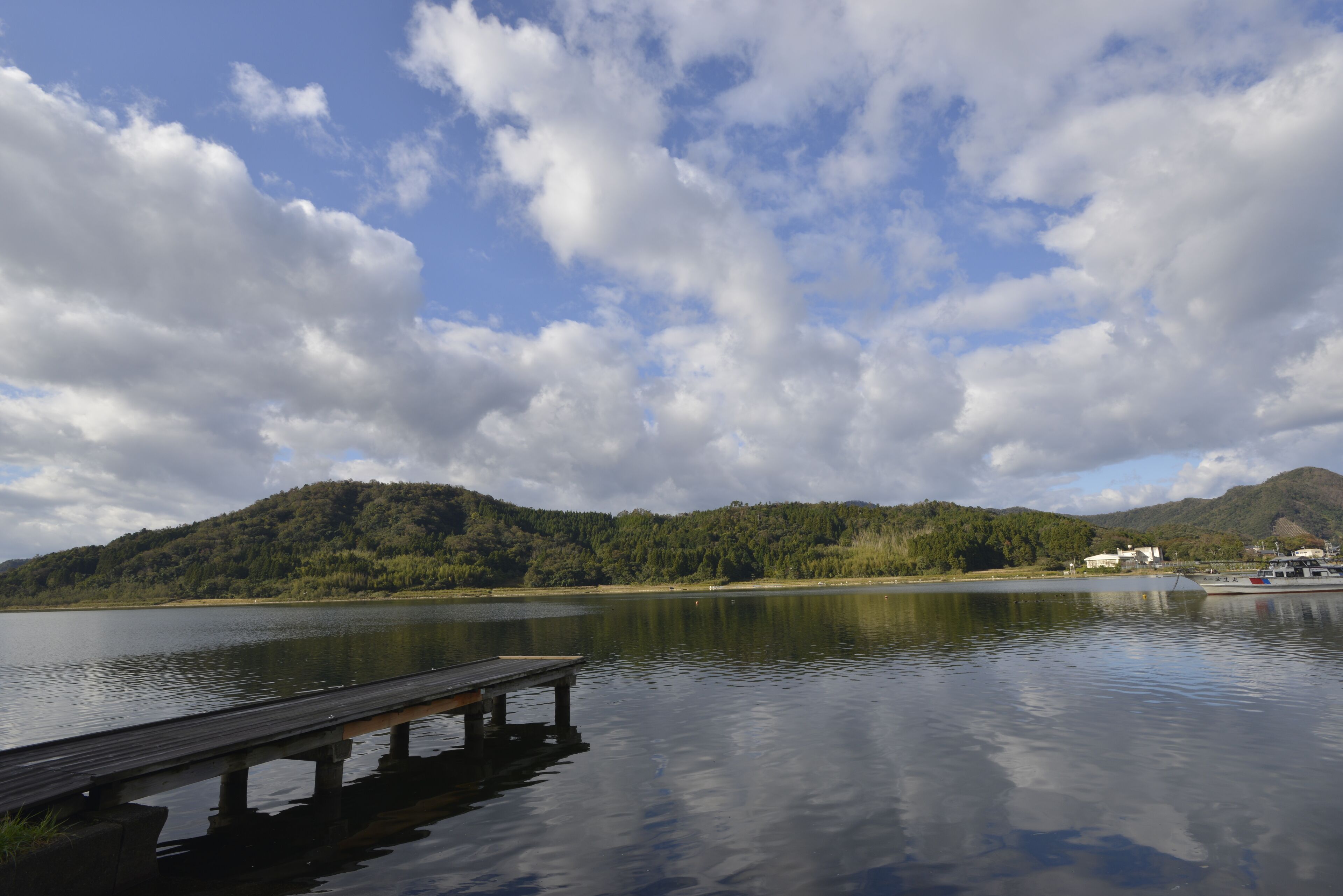 A calm lake with a wooden dock extending into the water, surrounded by lush green hills and a partly cloudy sky.