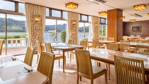Un restaurante con varias mesas y sillas, ventanas amplias con cortinas, y vistas a un lago y montañas.