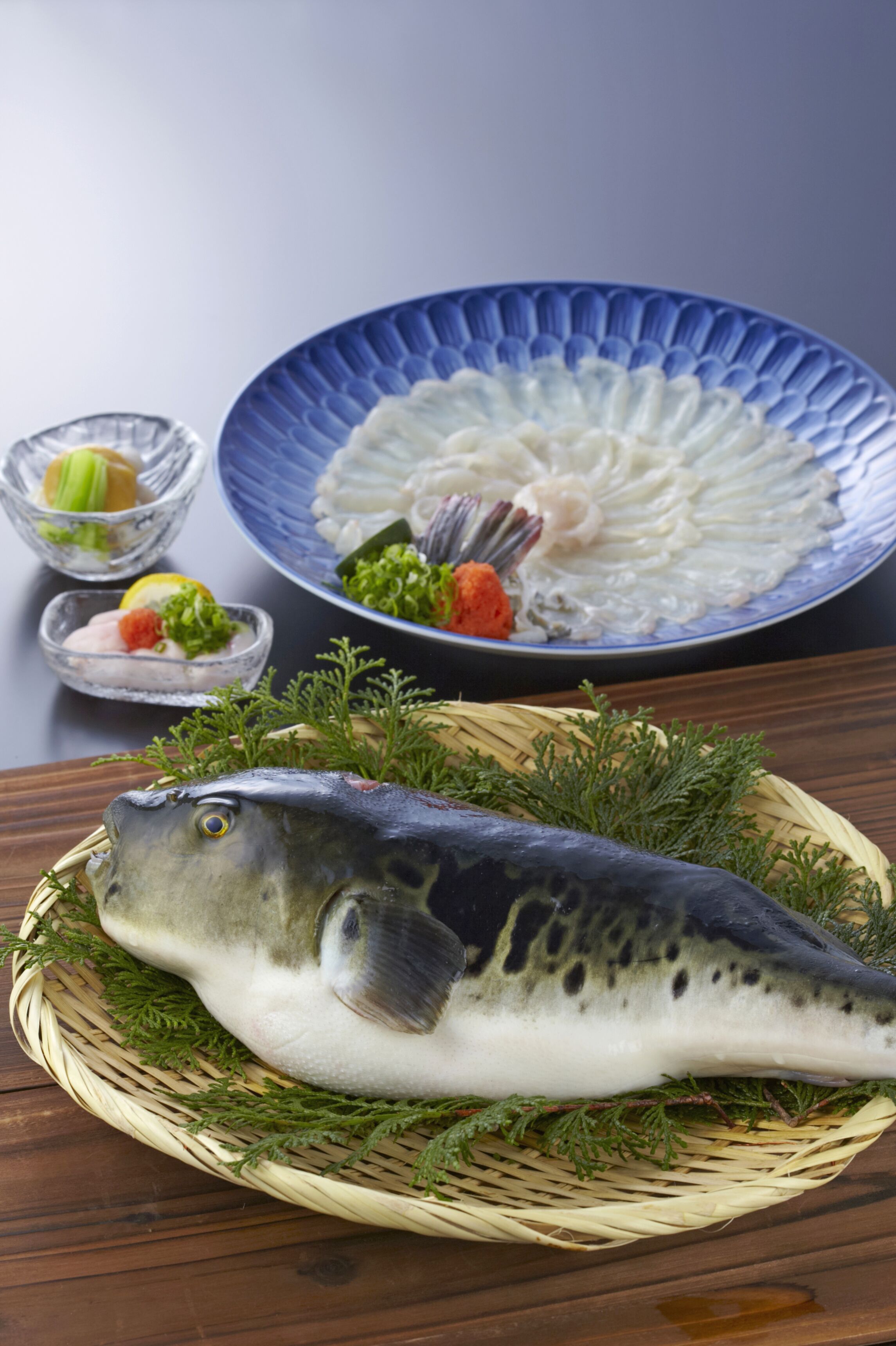 A fish on a woven basket with a blue plate of noodles and two small bowls of side dishes.
