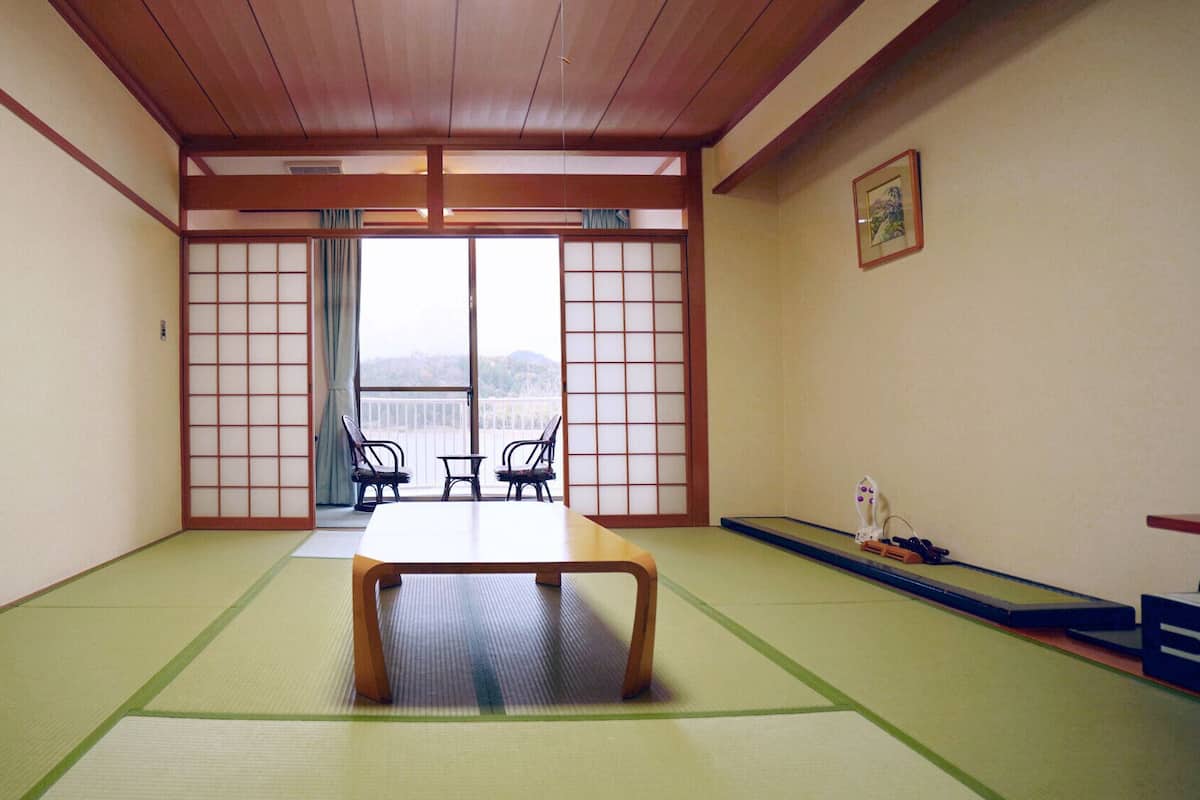 Une pièce avec un sol recouvert de tatamis, une petite table et des chaises, des portes coulissantes et une vue sur l’extérieur.