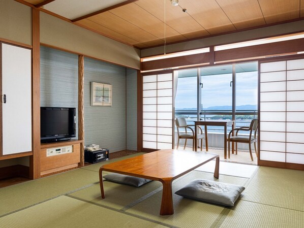 A traditional Japanese room with tatami flooring, a low wooden table, a television, and a view of a body of water.