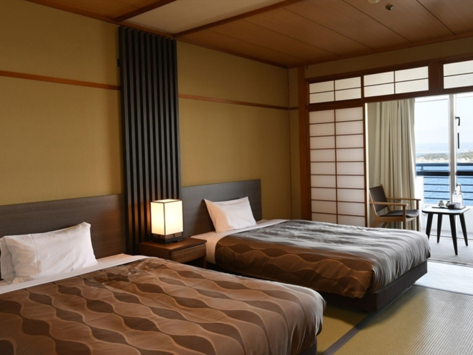 A hotel room with two beds, a nightstand, a lamp, a chair, and a view of the sea.