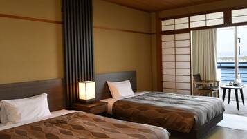 A hotel room with two beds, a nightstand, a lamp, a chair, and a view of the sea.