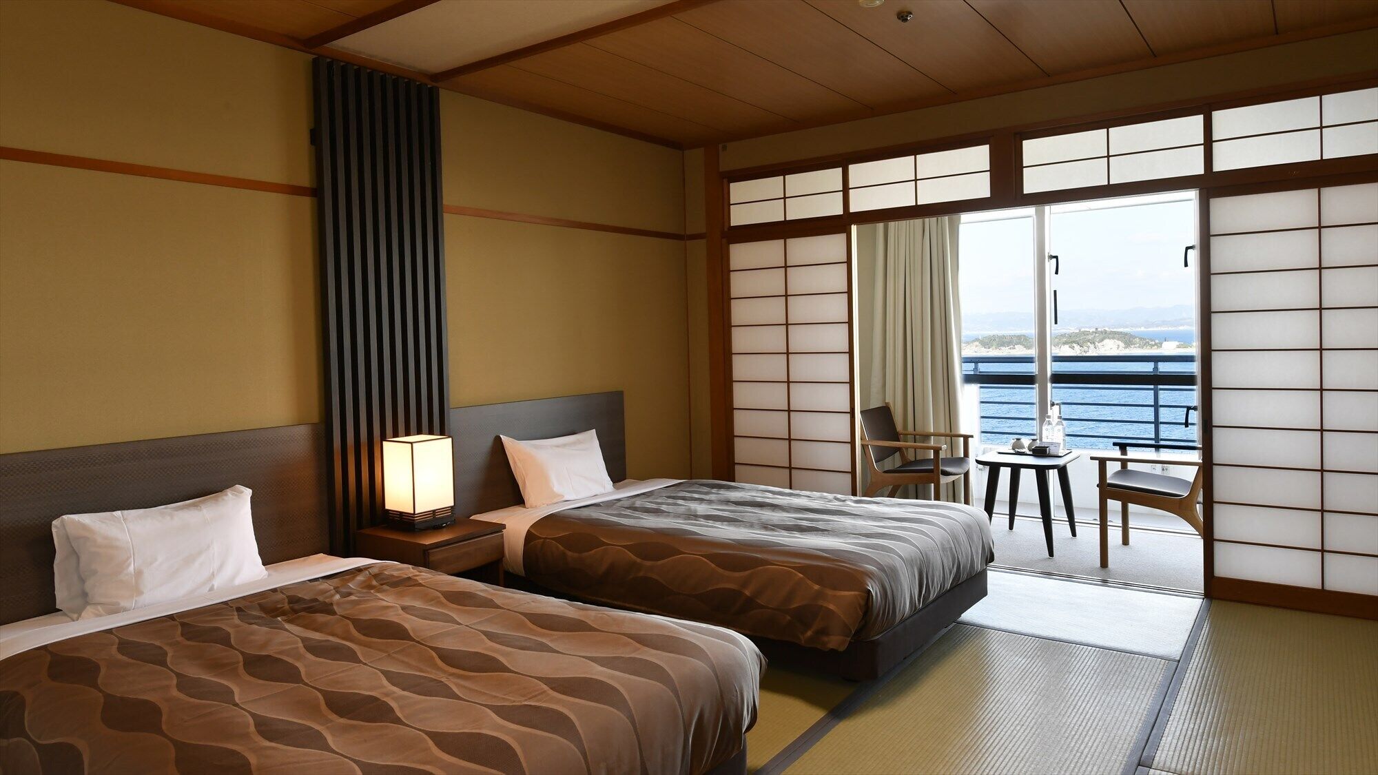 Une chambre d’hôtel avec deux lits, une table de chevet avec une lampe, une chaise, une petite table et une vue sur la mer.