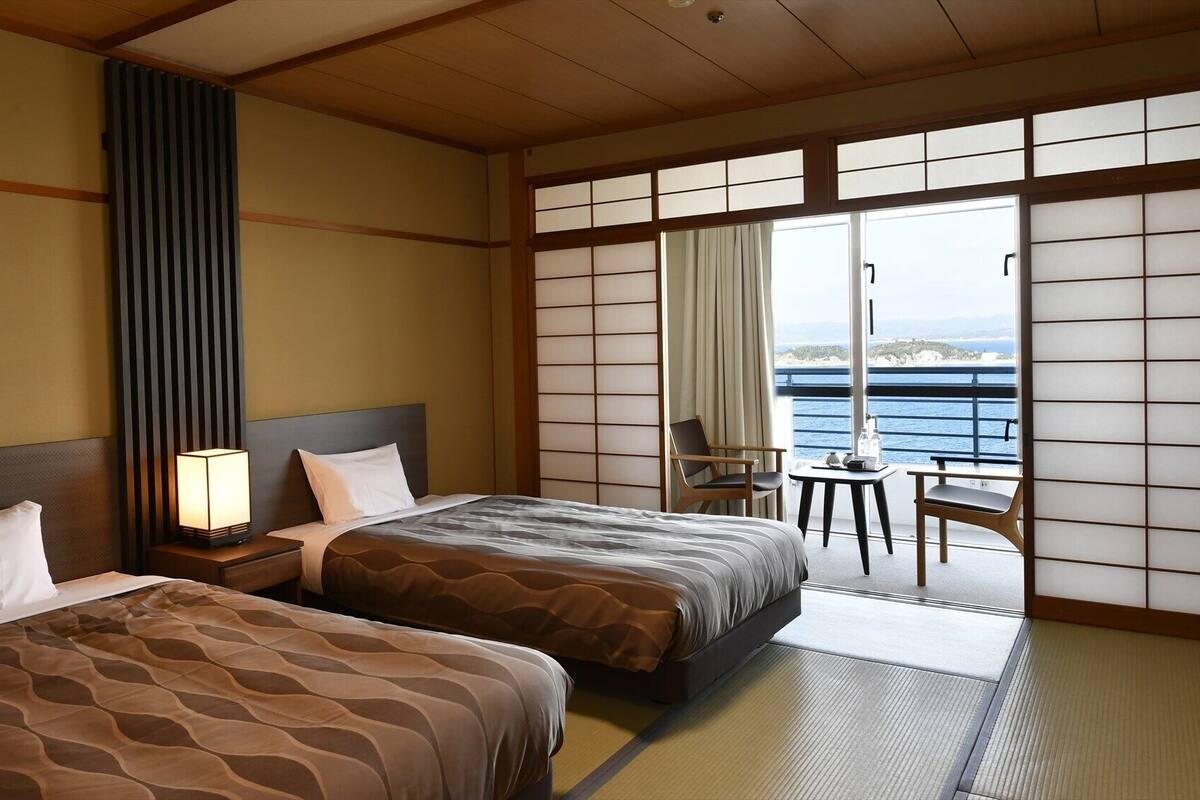 Habitación de hotel con dos camas, una mesita de noche con lámpara, una silla, una pequeña mesa y vista al mar.