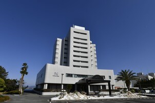 Un hotel moderno con una entrada destacada, varios pisos y una vista despejada del océano al fondo.
