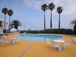 Una piscina con sillas y mesas blancas, rodeada de palmeras y un cielo despejado.