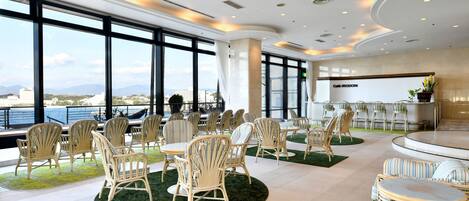 Amplio espacio interior con grandes ventanales que ofrecen vistas a la ciudad y las montañas, amoblado con sillas y mesas de mimbre, y una zona de bar con un letrero que dice "Café IRODORI".