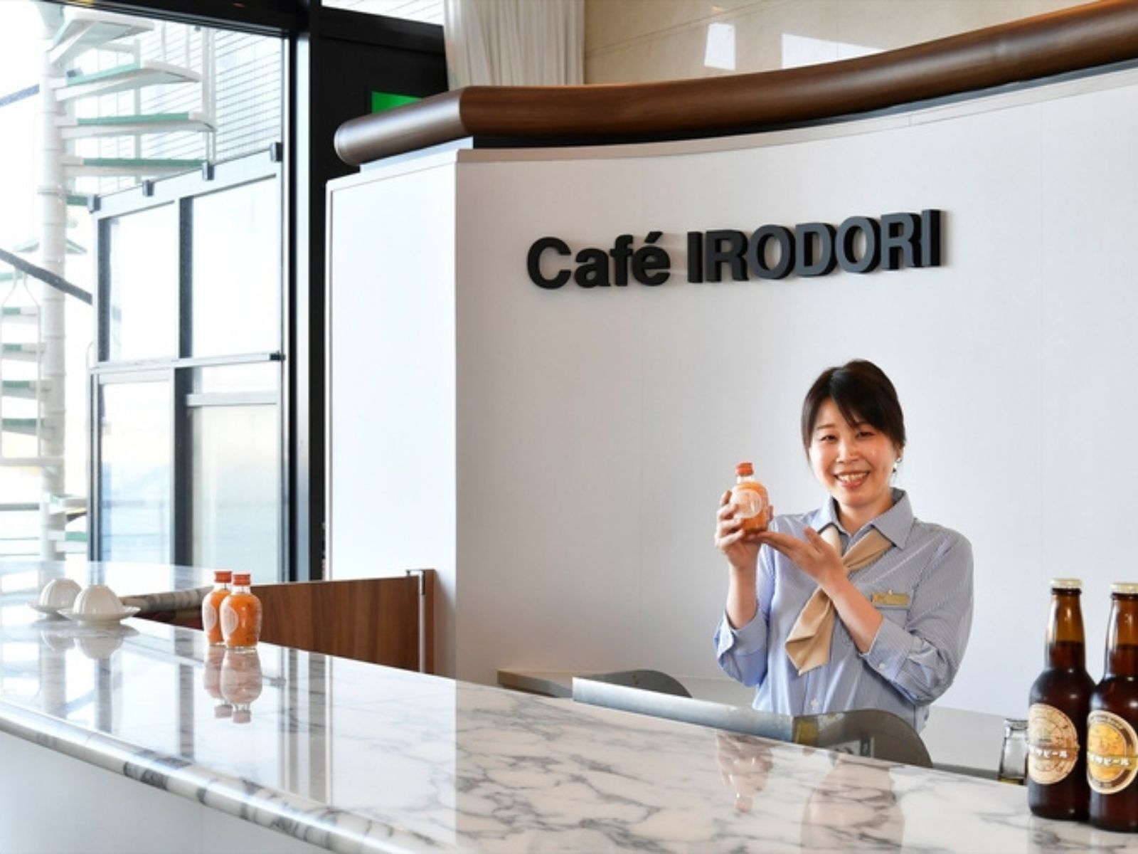 A person standing behind a counter with a sign reading 'Café IRODORI' and holding a bottle. There are two bottles on the counter.