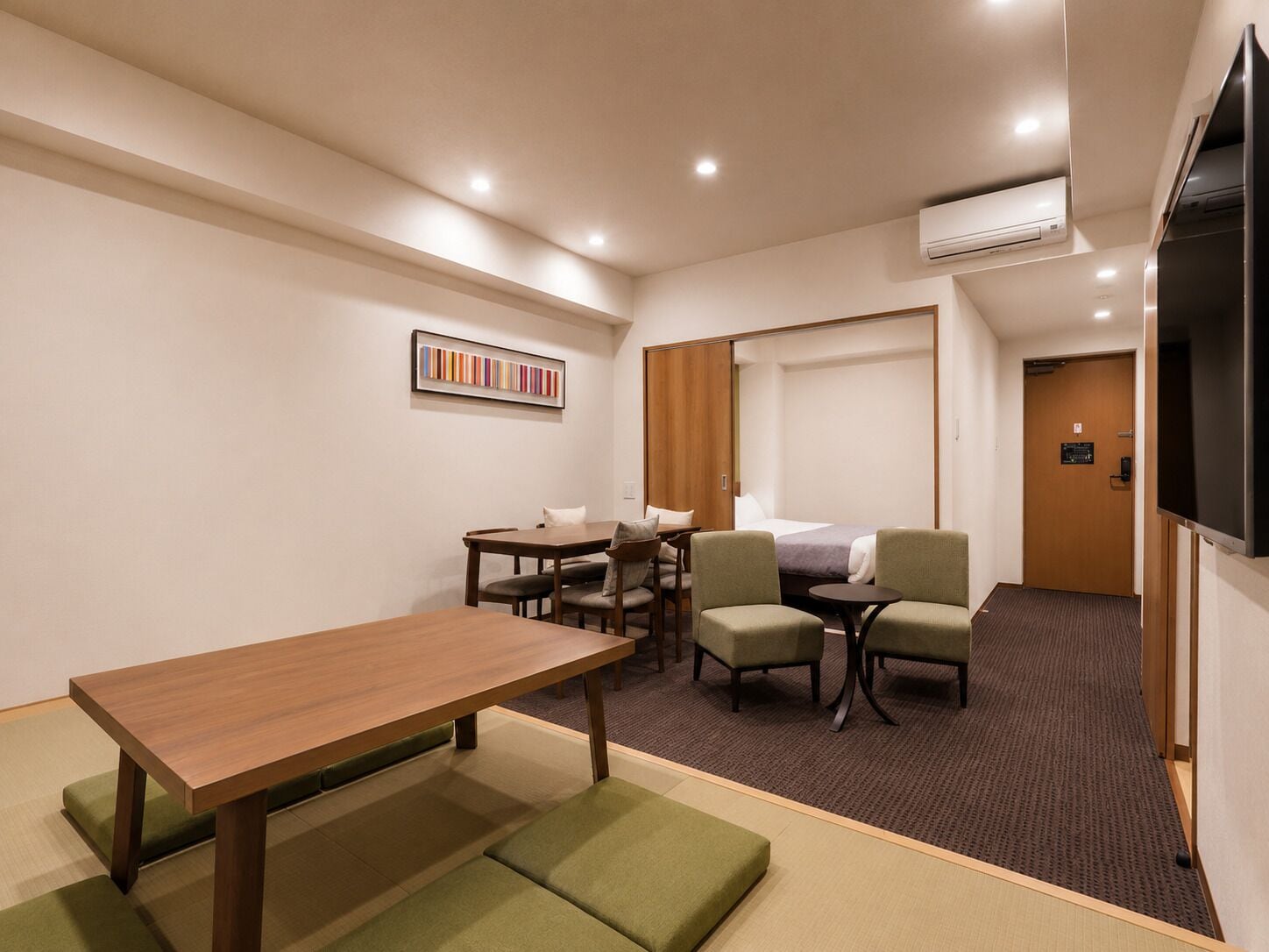 Een hotelkamer met een houten tafel, groene zittingen, een bed, een televisie en een airconditioning.