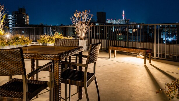 A rooftop terrace with a dining table and chairs, a bench, and a railing overlooking a cityscape at night. - Randor Residential Hotel Kyoto Suites (Kyoto)