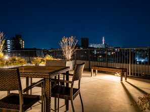 Una terraza en la azotea con mesa y sillas, un banco y una barandilla con vistas a un paisaje urbano nocturno.