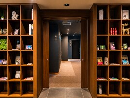 Um corredor com estantes de madeira em ambos os lados, contendo livros, objetos decorativos e uma placa indicando o caminho para o banheiro.