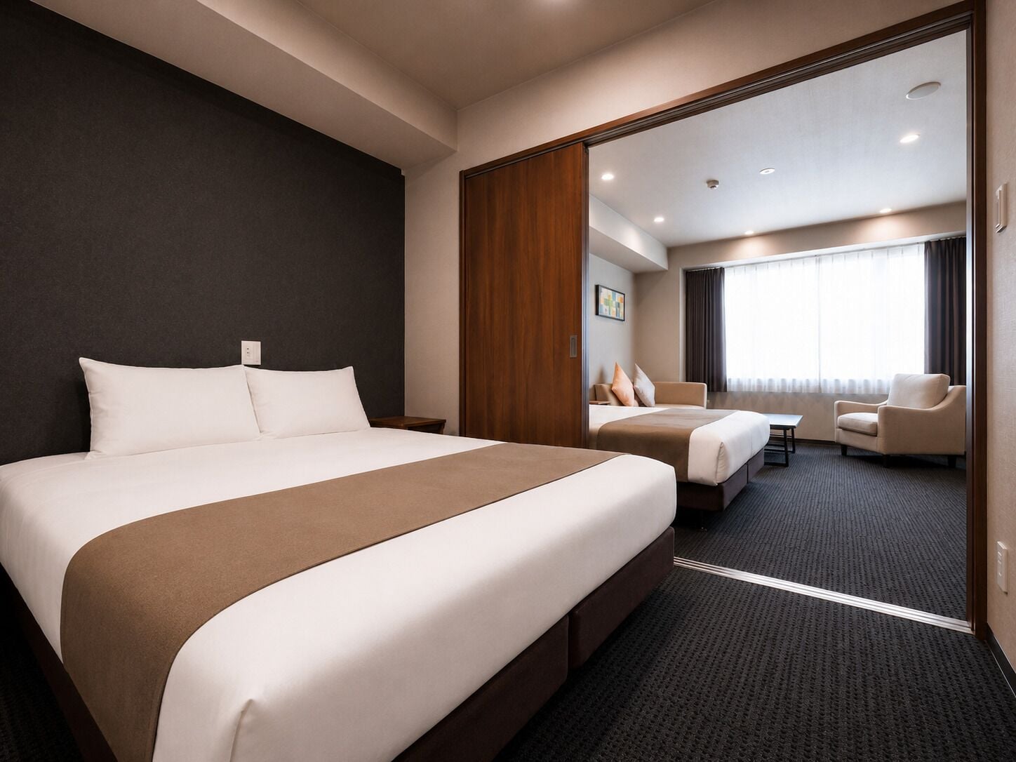 A hotel room with a large bed, a smaller bed, a desk, a chair, and a view of another room.