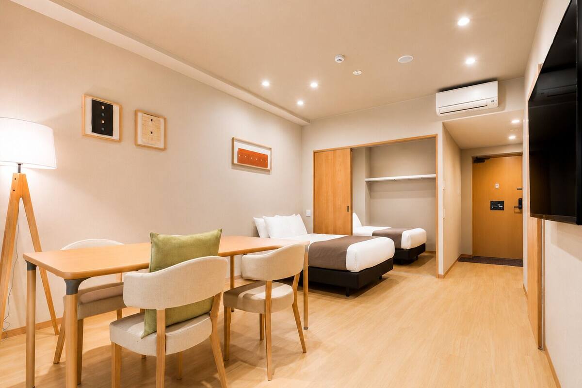A modern hotel room with a dining area, two beds, a TV, and an air conditioning unit.