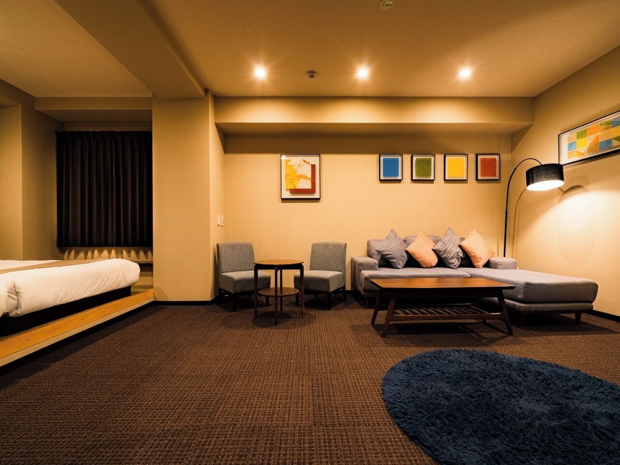 Habitación de hotel con cama, dos sillones, un sofá, una mesita, una lámpara y una alfombra azul.