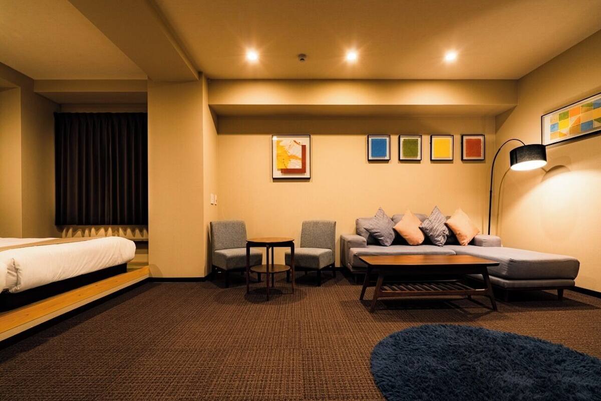 A hotel room with a bed, two armchairs, a sofa, a small table, a lamp, and a blue rug.