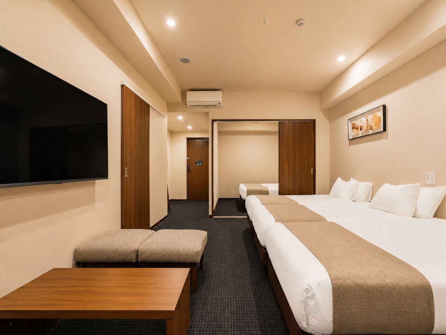 A hotel room with a TV, a desk, two beds, a sofa, and a hallway leading to another room.