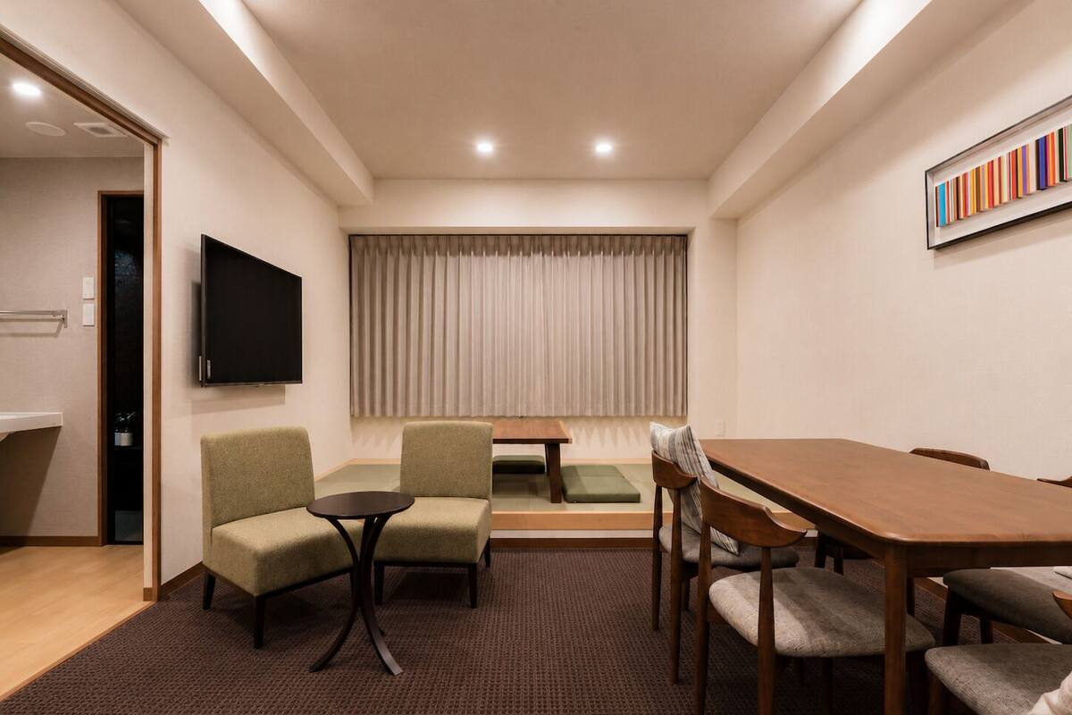 A hotel room with a wooden dining table, two green armchairs, a small round table, a TV mounted on the wall, and a window with curtains.