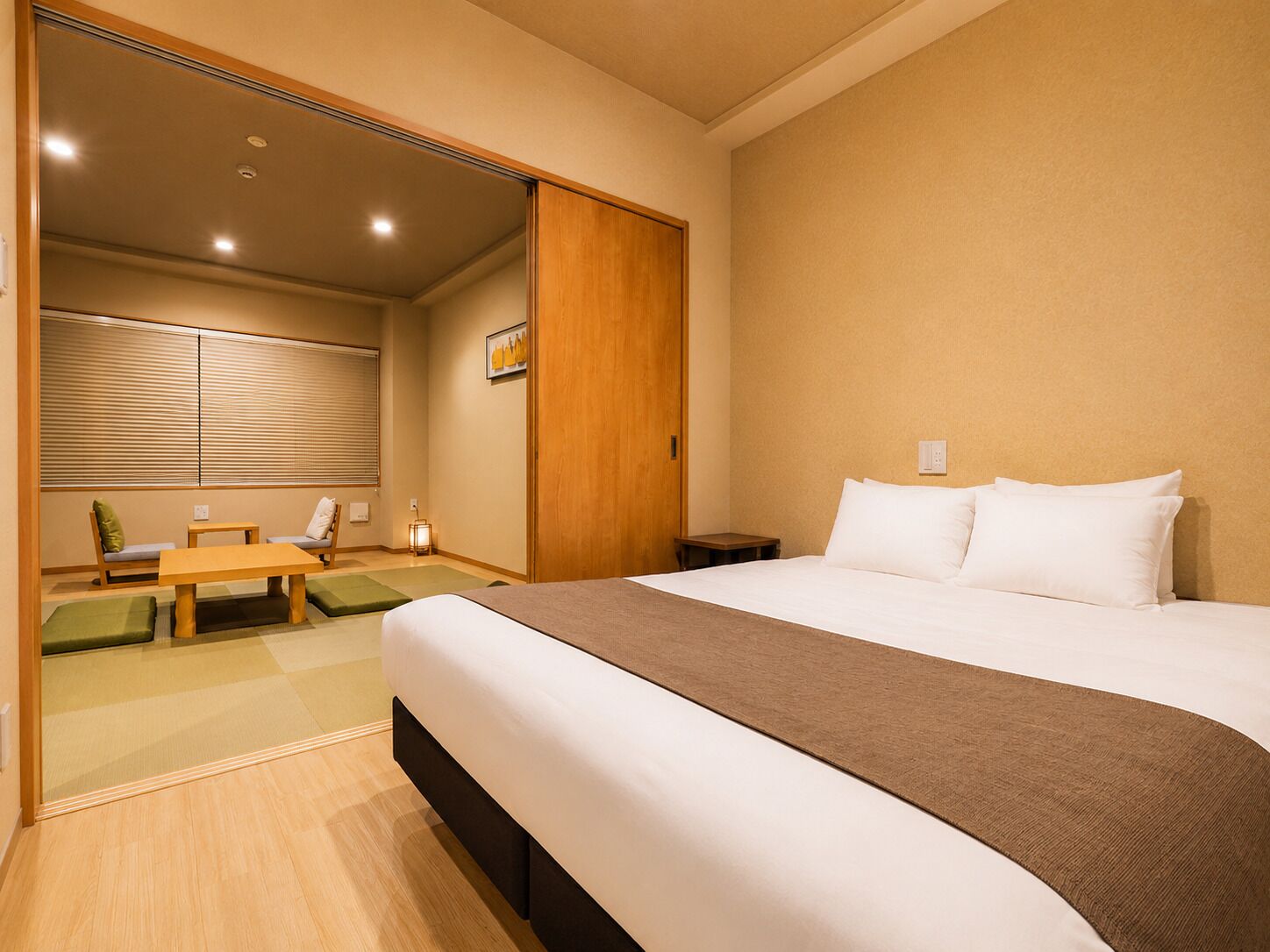 Une chambre d’hôtel comprenant un lit, un bureau, une chaise, une petite table, une fenêtre avec des stores et une armoire en bois.
