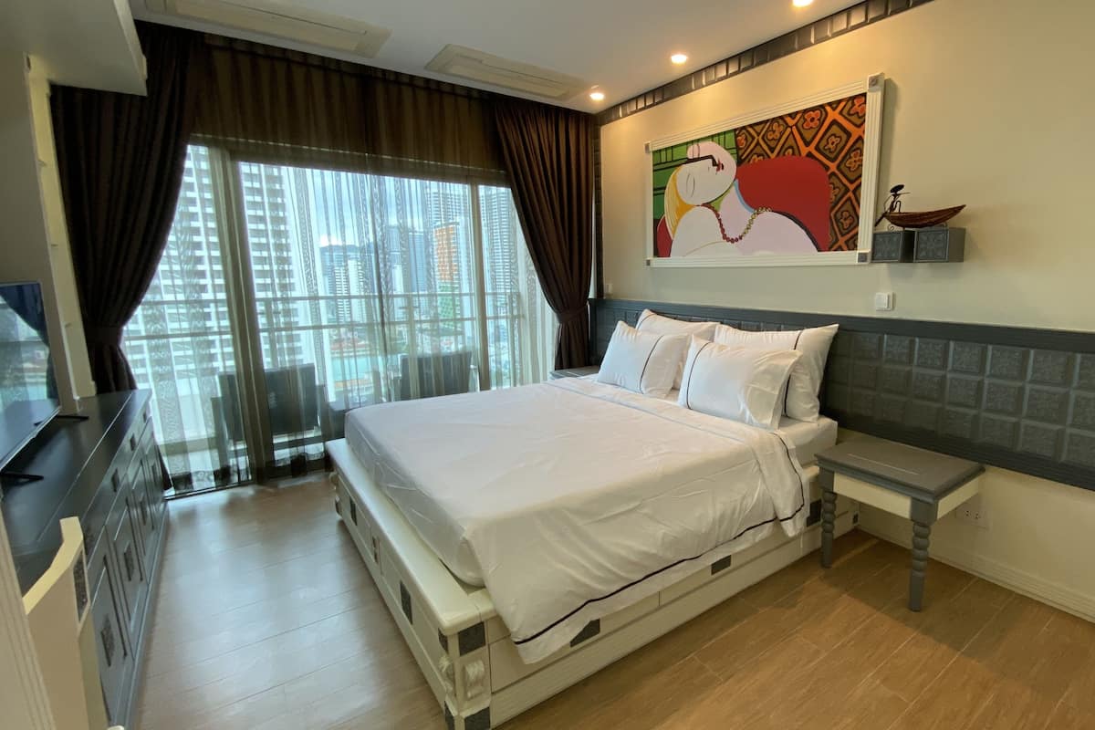 A hotel room with a large bed, a small bedside table, a TV, and a view of the city through the window.