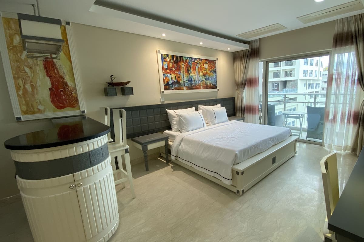 A modern hotel room with a large bed, a small desk, a chair, a trash bin, and a painting on the wall.